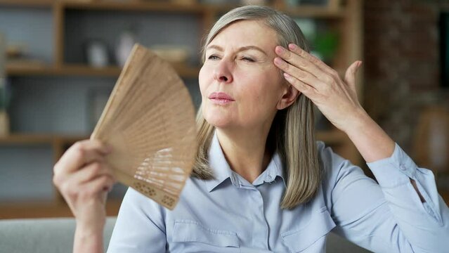 Senior female is hot while sitting on the sofa in the living room at home. Elderly woman waves a fan, wipes the sweat from her forehead with her hand. Air conditioner in apartment is broken. Close up