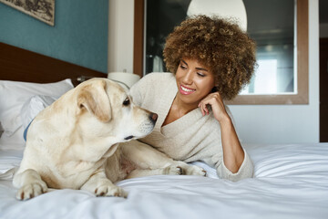 cheerful african american woman lying on bed and cuddling her labrador dog in pet friendly hotel