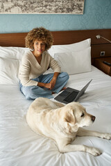 dreamy young african american woman sitting on bed near labrador and laptop in modern hotel room