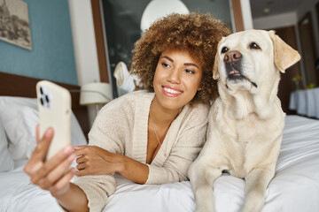 positive african american woman taking selfie with labrador on a bed in a pet-friendly hotel room