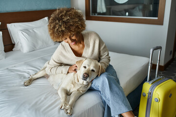 young and smiling african american woman cuddling labrador near luggage in pet-friendly hotel room