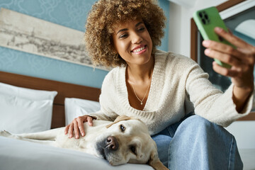 happy african american woman taking selfie with labrador on a bed in a pet-friendly hotel room
