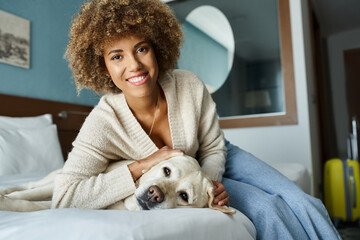 young and happy african american woman cuddling labrador on a bed in a pet-friendly hotel room