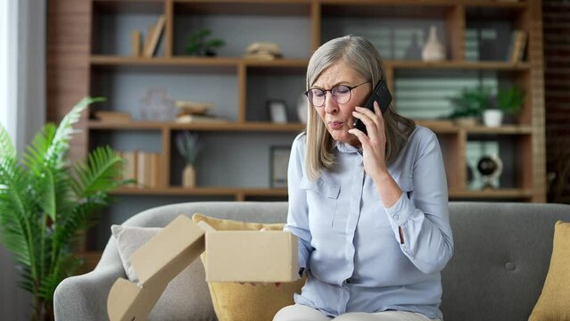 Unhappy senior female arguing on the phone after receiving a parcel with the wrong product at home. Elderly woman makes a bad review, she is dissatisfied with shopping in an online store. Damaged item