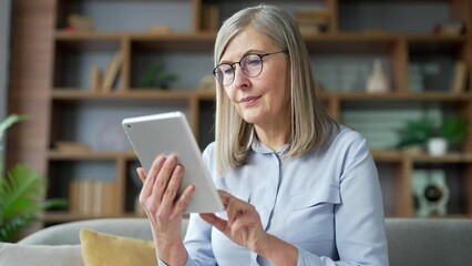 Senior woman with gray hair browsing digital tablet sitting on sofa in living room at home. Elderly female pensioner reads, writes messages, chats, does online shopping, uses an application. Close up - Powered by Adobe