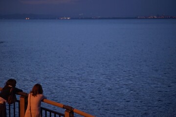 夕暮れの海を柵越しに眺める二人組の風景