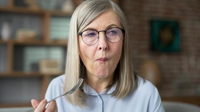 Happy senior gray haired female eating salad sitting on the sofa in living room at home. Smiling elderly retired woman enjoying a healthy meal, satisfied with a vegetarian food recipe diet. Close up