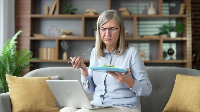 Tired of dieting for weight loss senior female with a plate of salad sitting on the sofa in the living room at home. Dissatisfied woman holds a fork in her hand, but does not want to eat healthy food
