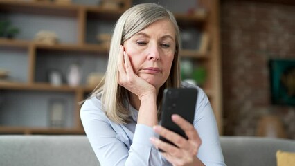 Lonely sad senior gray haired female bored using smartphone sitting on sofa in living room at home. Tired elderly retired woman is depressed and in a bad mood, browses social media on a mobile phone