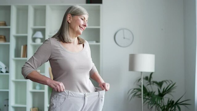 Happy senior gray haired female feels joy after losing weight. A smiling, satisfied middle aged woman tries on her big pants, which she wore before the diet while standing in the living room at home