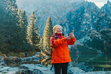 Rear shot of traveler 60s woman influencer in sportswear with backpack standing on the rocky shore and making selfie on smart phone. Taking photo of green hills and mountains on Morskie Oko lake