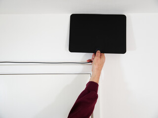 A man’s hand holding a modern, thin, lightweight, digital TV antenna on an interior room wall above a door, to find the best signal reception.