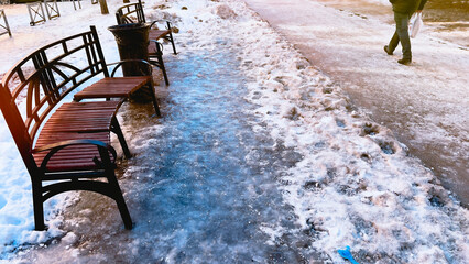Wooden benches on icy frozen ground and pavement in winter. Anonymous person walking along pavement...