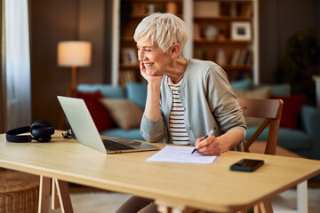 A cheerful senior adult woman working from home using laptop and writing reports