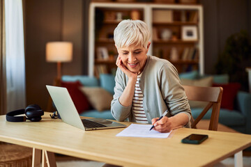 A smiling senior female accountant writing financial reports in front of a laptop in a home office