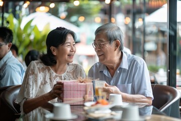 An Asian couple surprised an elderly father with a birthday gift at an outdoor cafe restaurant on their summer vacation. A celebration of father's day and a concept of elder care is part of the - obrazy, fototapety, plakaty