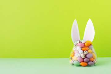Colorful candy jar decorated with bunny ears against green background, gifts for Easter