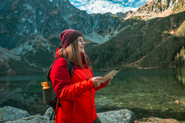 Shot of lost woman traveler with map and backpack relaxing outdoor with rocky on Morskie Oko lake. Woman traveler explorer with backpack hiking to the top of the mountain. Hiking travel and adventure - obrazy, fototapety, plakaty