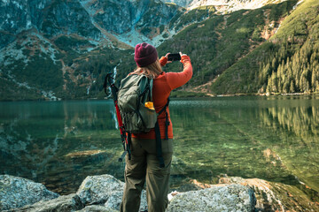 Rear shot of traveler woman in sportswear with backpack standing on the rocky shore and making selfie on smart phone. Taking photo of green hills and mountains on Morskie Oko lake. Hiking travel and - obrazy, fototapety, plakaty
