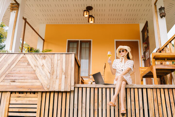 An Asian female hotel owner flies in from overseas to stay in a luxury wooden villa. Sitting and drinking wine in front of the room Working on a laptop, wearing a hat, dress, and sunglasses.