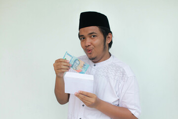 A young Asian muslim man holding an envelope and fifty thousand rupiah in cash with an expression...