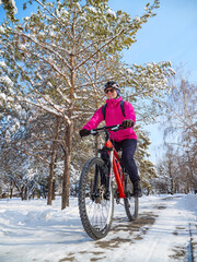 Winter cycling adventure. A girl in a pink jacket among snow-covered trees rides a bicycle along a path in the park