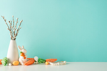 Fototapeta premium Easter display setup: Side view photo of a counter featuring a vase with pussy willow, a ceramic bunny figure, carrots for Easter bunny, assorted eggs, and beads on a pastel blue wall background