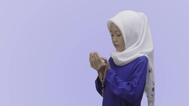 Muslim little girl in hijab praying with prayer beads while standing in blue muslim clothes