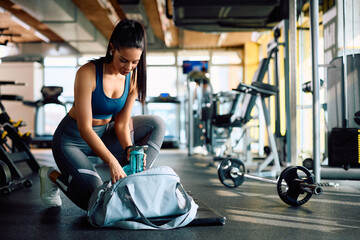 Young sportswoman taking water bottle from her sports bag in gym.