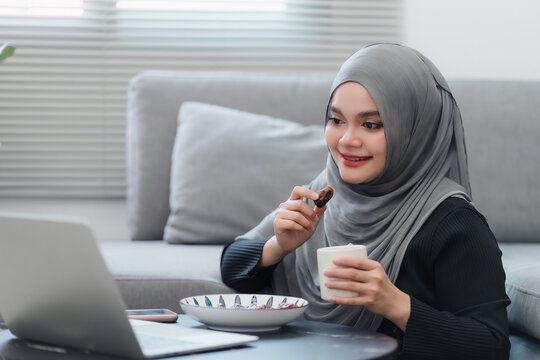 Cheerful Muslim woman in hijab eating dates fruit and enjoying a break at her laptop, in a comfortable home.