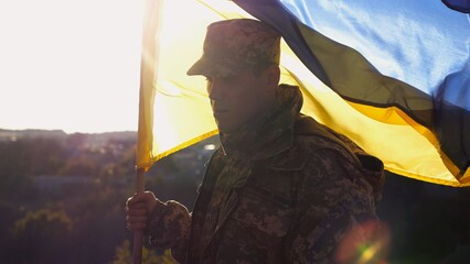 Soldier of ukrainian army stands with lifting blue-yellow banner at field. Male military in...