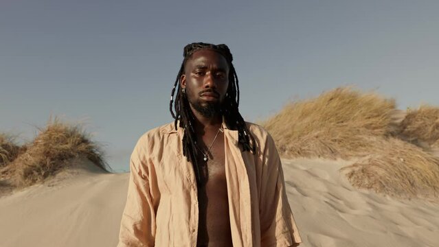 A confident man with fashionable dreadlocks standing in a serene desert setting, exuding a sense of freedom and style.