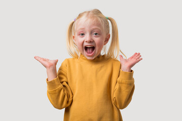 A cute little girl with blond hair in a yellow sweater stands with her mouth open and her hands raised.