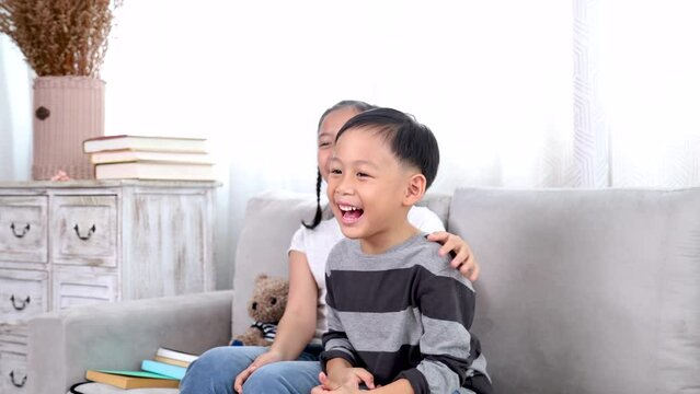 Asian siblings boy and girl share a laugh on a cozy sofa, happy children laugh bonding love relationship at home playtime, family lifestyle, enjoy carefree weekend, health care for kids concept