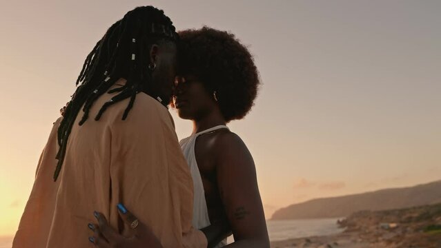 African American couple shares a tender embrace, bathed in the warmth of a serene sunset against a scenic outdoor landscape, symbolizing love and intimate connection.