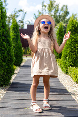 Summer portrait of a little girl in sunglasses, dress and straw hat. Childhood, outdoor leisure, vacations. Preschooler girl enjoying nature at country house.