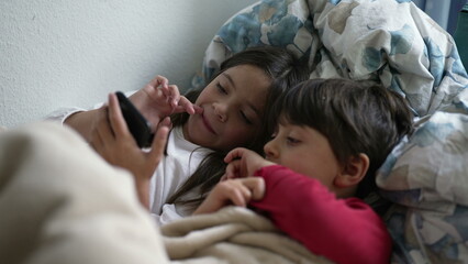 Children looking at cellphone device lying in bed under bedsheets, brother and sister absorbed by...