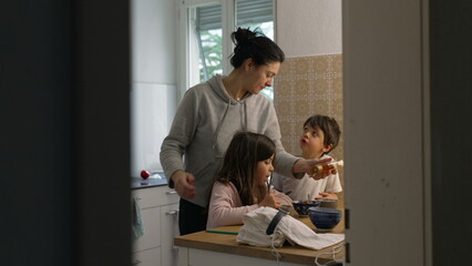 Candid domestic lifestyle scene of mother standing in kitchen with her two children preparing meal together. Small brother and sister interacting with parent while she pours honey into bowl