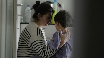 Caring mother consoling crying little son in candid kitchen scene. Mom embracing hurt child, candid...