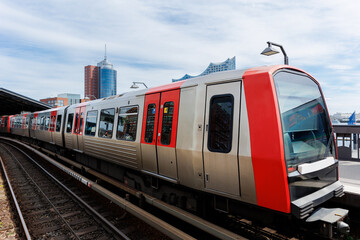 Fototapeta premium Hamburg Red Unmanned U-bahn Subway train on Baumwall station on Elevated Track with Hamburger Port Harbor Elbphilharmonie building background blue sky. Hanseatic city commute transport scenic view