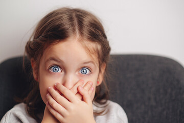 Little girl covers mouth with hand. Sad offended child girl sitting at sofa at home. Child abuse.