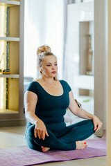 Vertical shot of full-figured mature middle age woman sitting on mat in lotus pose. Calm old lady practicing techniques and yoga indoors.
