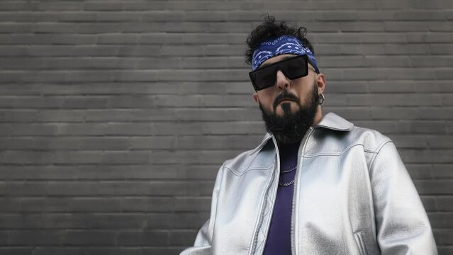 A cool bearded man with stylish sunglasses enjoys dancing outdoors in front of a grey wall, exuding a vibe of freedom and joy.