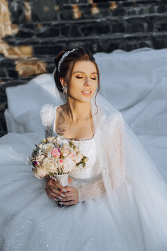 The brunette bride, smiling sincerely, sits on the ground in her unfolded dress and holds a bouquet. Wedding ceremony. Sunny day. Beautiful hair and makeup