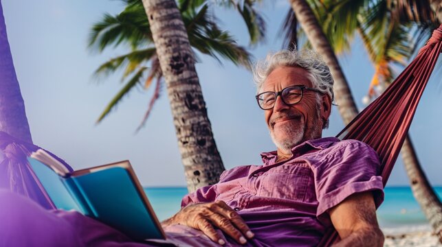 Senior man smiling contentedly as he relaxes in a hammock and reads a book under the shade of palm trees on a tranquil beach