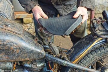 one  old rubber black saddle in the hand  near  the iron rusty  frame of a black dirty retro...