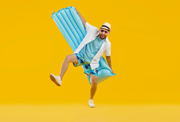 Funny overjoyed excited man wearing beach clothes with open mouth holding inflatable mattress isolated on a studio yellow background. Happy tourist is going on summer holiday trip. Vacation concept. - obrazy, fototapety, plakaty