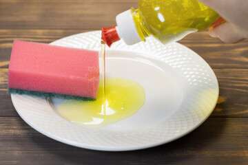 Man is pouring liquid yellow washer gel into sponge for washing dishes on the white plate. Soap for kitchen house cleaning.