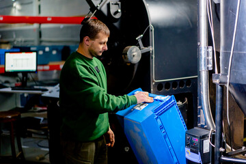 worker at a coffee factory pours green coffee from a blue box int a coffee roasting mechanism