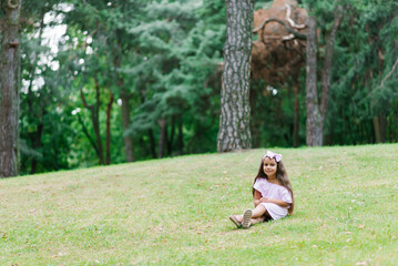 A happy little baby girl in a beautiful pink dress is sitting in the park on the grass and looking at the camera.